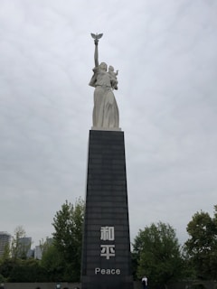 A tall statue stands atop a large pedestal, featuring a figure holding a dove aloft with one arm while supporting a child with the other arm. The pedestal displays the word 'Peace' and its translation in another language. Surrounding the monument are trees and some buildings are visible in the background under an overcast sky.