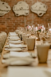 A long dining table elegantly set with white napkins, plates, and clear wine glasses, softly lit by candles. The brick wall in the background features abstract artwork, creating a cozy and intimate atmosphere.