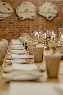 A long dining table elegantly set with white napkins, plates, and clear wine glasses, softly lit by candles. The brick wall in the background features abstract artwork, creating a cozy and intimate atmosphere.