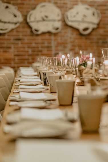 A long dining table elegantly set with white napkins, plates, and clear wine glasses, softly lit by candles. The brick wall in the background features abstract artwork, creating a cozy and intimate atmosphere.