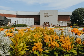 A well-maintained garden filled with vibrant yellow and orange flowers, leading up to a large modern building with a white and brown facade. The building has an inscription in Russian on the main entrance wall. There are several people walking and standing around the garden area, and a sculpture of a tank is visible on the left side of the image.