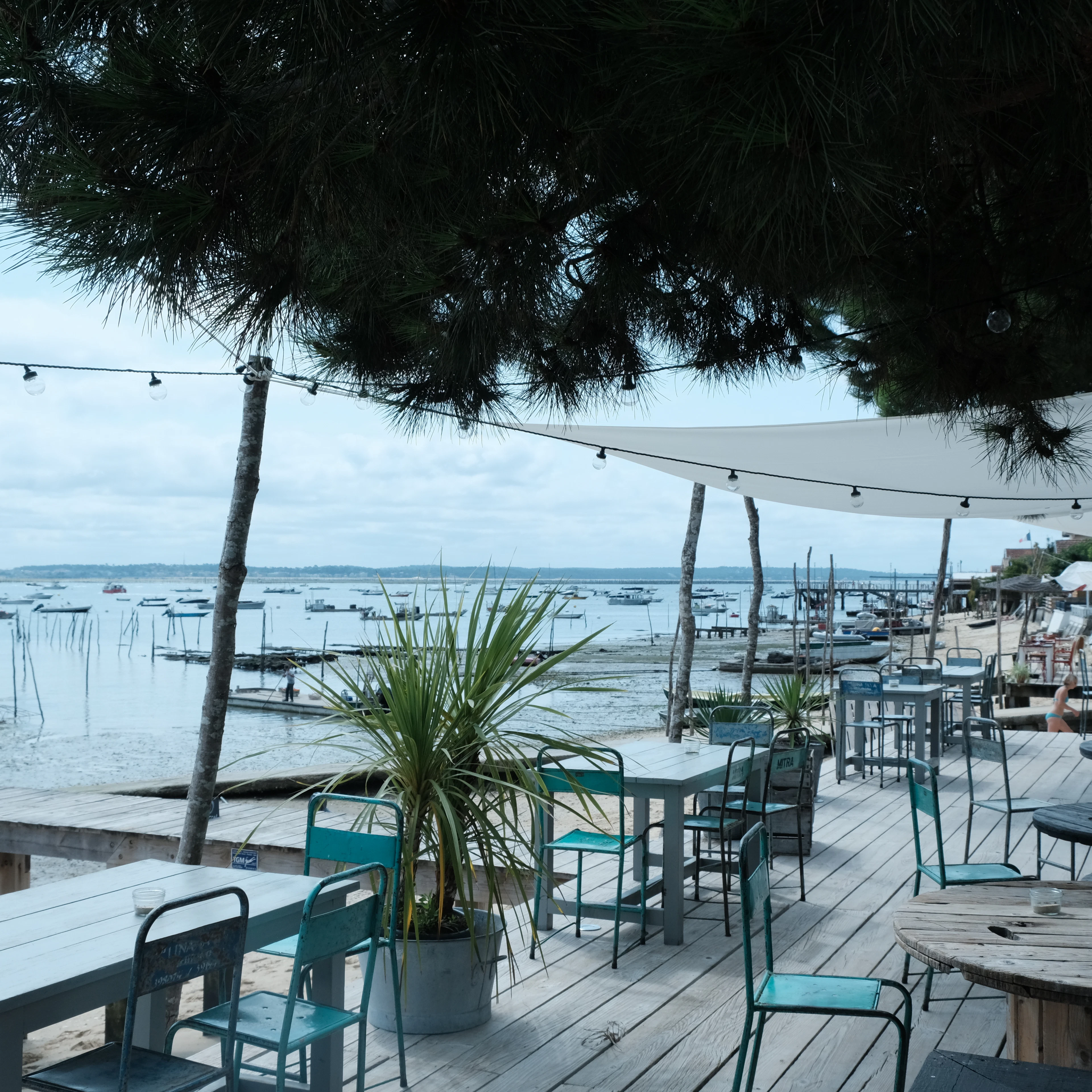Weathered teal chairs and long wooden tables on the deck of an oyster shack at Cap Ferret, string lights hung between pines, looking out over the oyster beds and moored boats of the Bassin d'Arcachon.