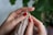 Hands with red nail polish are actively knitting with beige yarn. The intricacy of the knitting is visible, with the yarn wrapped around wooden knitting needles. A leafy green plant serves as a backdrop, providing a natural contrast to the focused crafting activity.