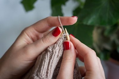 Hands with red nail polish are actively knitting with beige yarn. The intricacy of the knitting is visible, with the yarn wrapped around wooden knitting needles. A leafy green plant serves as a backdrop, providing a natural contrast to the focused crafting activity.