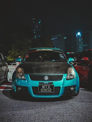 A modified Suzuki car is parked in an urban area at night. The car features a bright turquoise front bumper and black hood with various decals on the windshield. Its headlights are on, illuminating the parking space. Surrounding the car are other vehicles in a dimly lit environment, with high-rise buildings visible in the background.