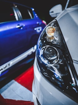A close-up view of the front headlight of a white car, with reflections and details of the light elements. In the background, part of a blue car is visible with a focus on its reflective side surfaces. The scene is illuminated with artificial lighting, highlighting the textures and surfaces of the cars.
