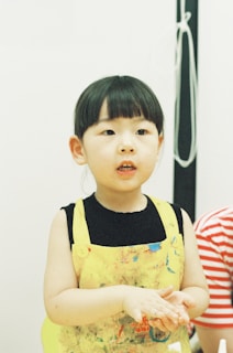 A young child wearing a colorful apron, possibly with paint stains, stands with hands together. The background is simple, with a hint of a vertical black element and a coil of white cord.