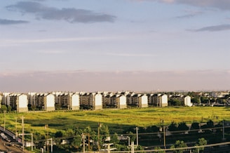 A row of identical, multi-story residential buildings stretches across a lush, open green field. The buildings are uniformly aligned and the scene is lit by a warm, soft lighting, suggesting early morning or late afternoon. In the foreground, there is a road with some traffic infrastructure, including power lines and a few vehicles. The sky above is expansive and mostly clear, with some light clouds.