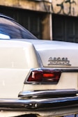 A close-up view of the rear corner of a vintage white car featuring a badge that reads '280 SE AUTOMATIC'. The taillight is visible, along with a chrome bumper showing some wear. The background consists of a blurred wall with dark windows and graffiti.