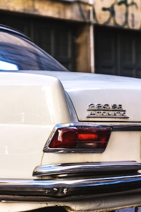 A close-up view of the rear corner of a vintage white car featuring a badge that reads '280 SE AUTOMATIC'. The taillight is visible, along with a chrome bumper showing some wear. The background consists of a blurred wall with dark windows and graffiti.