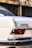 A close-up view of the rear corner of a vintage white car featuring a badge that reads '280 SE AUTOMATIC'. The taillight is visible, along with a chrome bumper showing some wear. The background consists of a blurred wall with dark windows and graffiti.
