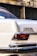 A close-up view of the rear corner of a vintage white car featuring a badge that reads '280 SE AUTOMATIC'. The taillight is visible, along with a chrome bumper showing some wear. The background consists of a blurred wall with dark windows and graffiti.