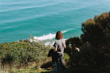 A woman standing on a cliff overlooking a vast turquoise ocean, her hair catching the breeze.