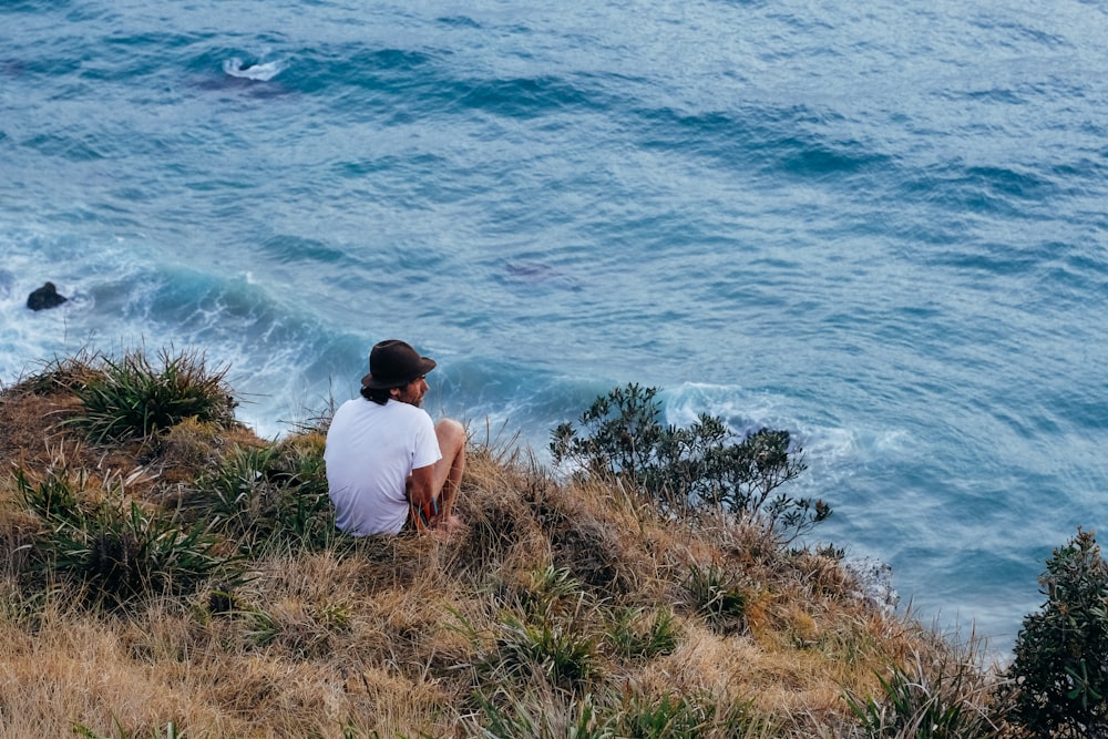 man in white t-shirt sitting on green grass near body of water during daytime