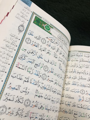 A close-up view of an open book featuring an ornate and colorful page with Arabic calligraphy. The text includes decorative borders and symbols with different colored fonts for emphasis. The page layout is intricate and aesthetically pleasing.