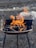 A metal grill with dark coals ignited by vibrant orange flames, set against a rough stone or gravel background.