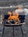 A metal grill with dark coals ignited by vibrant orange flames, set against a rough stone or gravel background.