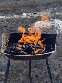 A metal grill with dark coals ignited by vibrant orange flames, set against a rough stone or gravel background.