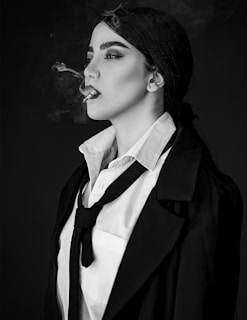 A person in a formal outfit with a tie and coat is smoking. Smoke trails from the cigarette, adding a dramatic element. The composition is in black and white, highlighting the contrast and shadows on the face and clothing.