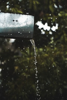 A metal gutter with water flowing from it in a smooth stream, surrounded by droplets. The background consists of blurred green foliage, suggesting a natural setting.