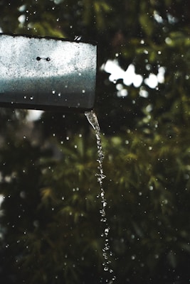 A metal gutter with water flowing from it in a smooth stream, surrounded by droplets. The background consists of blurred green foliage, suggesting a natural setting.