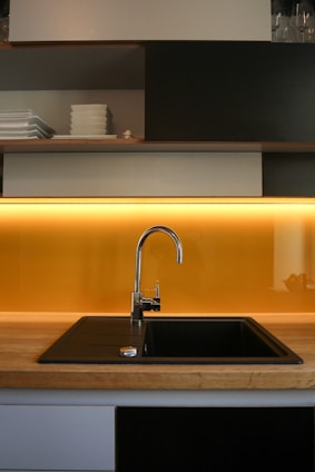 A modern kitchen features clean lines with open shelving filled with neatly stacked white dishes. The focal point is the sleek, metal faucet centered above a dark, square sink set into a wooden countertop. A warm, yellow backlighting adds a cozy, inviting glow to the minimalist design.