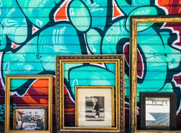 Three ornate picture frames are positioned against a vibrant graffitied wall. Within the frames, there is a painting depicting an outdoor scene, a black and white photograph of a person, and a reflective mirror. The graffiti features abstract shapes and bold colors.