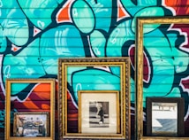 Three ornate picture frames are positioned against a vibrant graffitied wall. Within the frames, there is a painting depicting an outdoor scene, a black and white photograph of a person, and a reflective mirror. The graffiti features abstract shapes and bold colors.