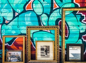 Three ornate picture frames are positioned against a vibrant graffitied wall. Within the frames, there is a painting depicting an outdoor scene, a black and white photograph of a person, and a reflective mirror. The graffiti features abstract shapes and bold colors.
