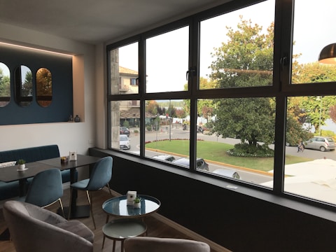A modern interior with a large window providing a view of a street scene outside. The room features stylish furniture including a black table, blue upholstered chairs, and wall decor with mirrors. Outside, trees and a parked car are visible, along with a few people walking.