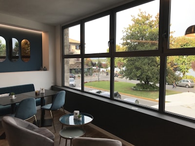 A modern interior with a large window providing a view of a street scene outside. The room features stylish furniture including a black table, blue upholstered chairs, and wall decor with mirrors. Outside, trees and a parked car are visible, along with a few people walking.