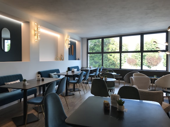A modern cafe interior with sleek, dark tables and teal upholstered seating. The space is well-lit by natural light from large windows overlooking greenery. The walls feature decorative lighting fixtures and small vases as accents. Minimalist decor creates a calm, inviting atmosphere.