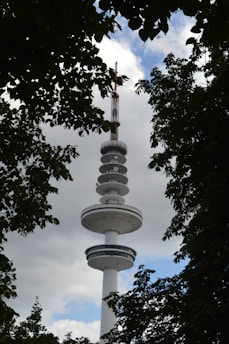 A tall communication tower with multiple disk-like platforms surrounded by lush green leaves. The top of the tower is visible against a partly cloudy sky with patches of blue.