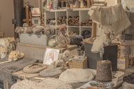 A cozy and rustic shop display featuring a variety of yarns and woven textiles is organized neatly. The shelves are filled with skeins of yarn in different shades, while woven blankets and shawls are draped for presentation. A small basket contains folded textiles, and knitting tools are laid out on the surface. Soft lighting enhances the warmth of the natural fibers, adding a welcoming atmosphere.
