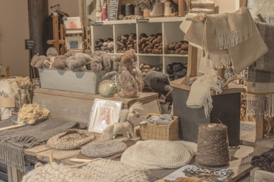 A cozy and rustic shop display featuring a variety of yarns and woven textiles is organized neatly. The shelves are filled with skeins of yarn in different shades, while woven blankets and shawls are draped for presentation. A small basket contains folded textiles, and knitting tools are laid out on the surface. Soft lighting enhances the warmth of the natural fibers, adding a welcoming atmosphere.