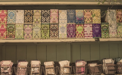 A selection of Pukka tea boxes arranged neatly on wooden shelves. Each box features vibrant and diverse artwork, showcasing various flavors such as peppermint, night time, and elderberry. Below the tea boxes, wire baskets hold additional packaged products.
