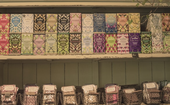 A selection of Pukka tea boxes arranged neatly on wooden shelves. Each box features vibrant and diverse artwork, showcasing various flavors such as peppermint, night time, and elderberry. Below the tea boxes, wire baskets hold additional packaged products.