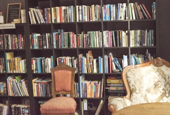 A well-stocked bookcase fills the background, with shelves packed with various books. In front of the shelves, there are two chairs: one upholstered in an ornate, floral pattern, and the other in a simple, reddish fabric. A small table is visible in the foreground. The lighting creates a cozy, warm atmosphere, suggesting a quiet and inviting reading space.