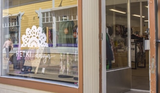 A store front with a large glass window displaying the logo and name 'HETKI design' in white lettering. Inside the store, various clothing items are visible on racks and shelves. The exterior features a yellow wooden building with white trim and some reflective surfaces.