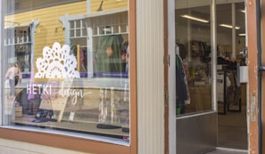 A store front with a large glass window displaying the logo and name 'HETKI design' in white lettering. Inside the store, various clothing items are visible on racks and shelves. The exterior features a yellow wooden building with white trim and some reflective surfaces.