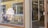 A store front with a large glass window displaying the logo and name 'HETKI design' in white lettering. Inside the store, various clothing items are visible on racks and shelves. The exterior features a yellow wooden building with white trim and some reflective surfaces.