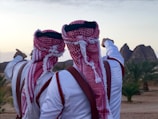 Two people wearing traditional red and white keffiyehs and robes stand side by side, pointing towards distant rock formations. The scene includes palm trees and a desert landscape under a slightly cloudy sky.