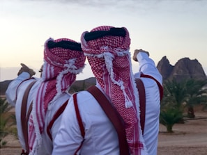 Two people wearing traditional red and white keffiyehs and robes stand side by side, pointing towards distant rock formations. The scene includes palm trees and a desert landscape under a slightly cloudy sky.