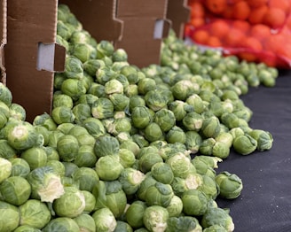 A large pile of fresh green Brussels sprouts is displayed on a black surface, with some of them spilling out of open cardboard boxes. In the background, there is a glimpse of a red mesh bag containing numerous oranges.