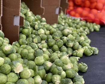 A large pile of fresh green Brussels sprouts is displayed on a black surface, with some of them spilling out of open cardboard boxes. In the background, there is a glimpse of a red mesh bag containing numerous oranges.