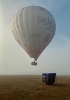 A hot air balloon with branding featuring skydiving images hovers above a foggy open field. The balloon is mostly white with red accents and text. It ascends above the grassy terrain, where there is a covered object nearby.