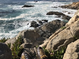 A pair of hiking boots positioned on rocky terrain overlooks a rugged coastline. Waves crash against the rocks, creating white foam that contrasts with the deep blue of the ocean. Sparse green vegetation grows between the rocks.