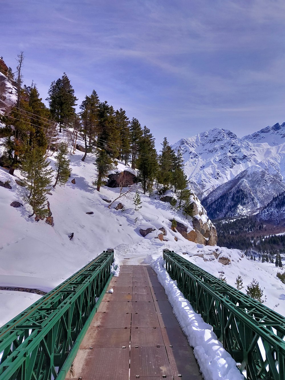 昼間の雪山に架かる緑色の金属橋