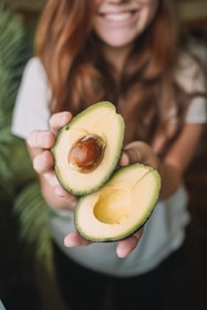 A person with long brown hair holds two halves of a ripe avocado in their hands. The avocado's flesh is light green and creamy, with one half containing the large seed. The background features blurred greenery and the person is smiling, creating a pleasant and fresh atmosphere.