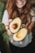 A person with long brown hair holds two halves of a ripe avocado in their hands. The avocado's flesh is light green and creamy, with one half containing the large seed. The background features blurred greenery and the person is smiling, creating a pleasant and fresh atmosphere.